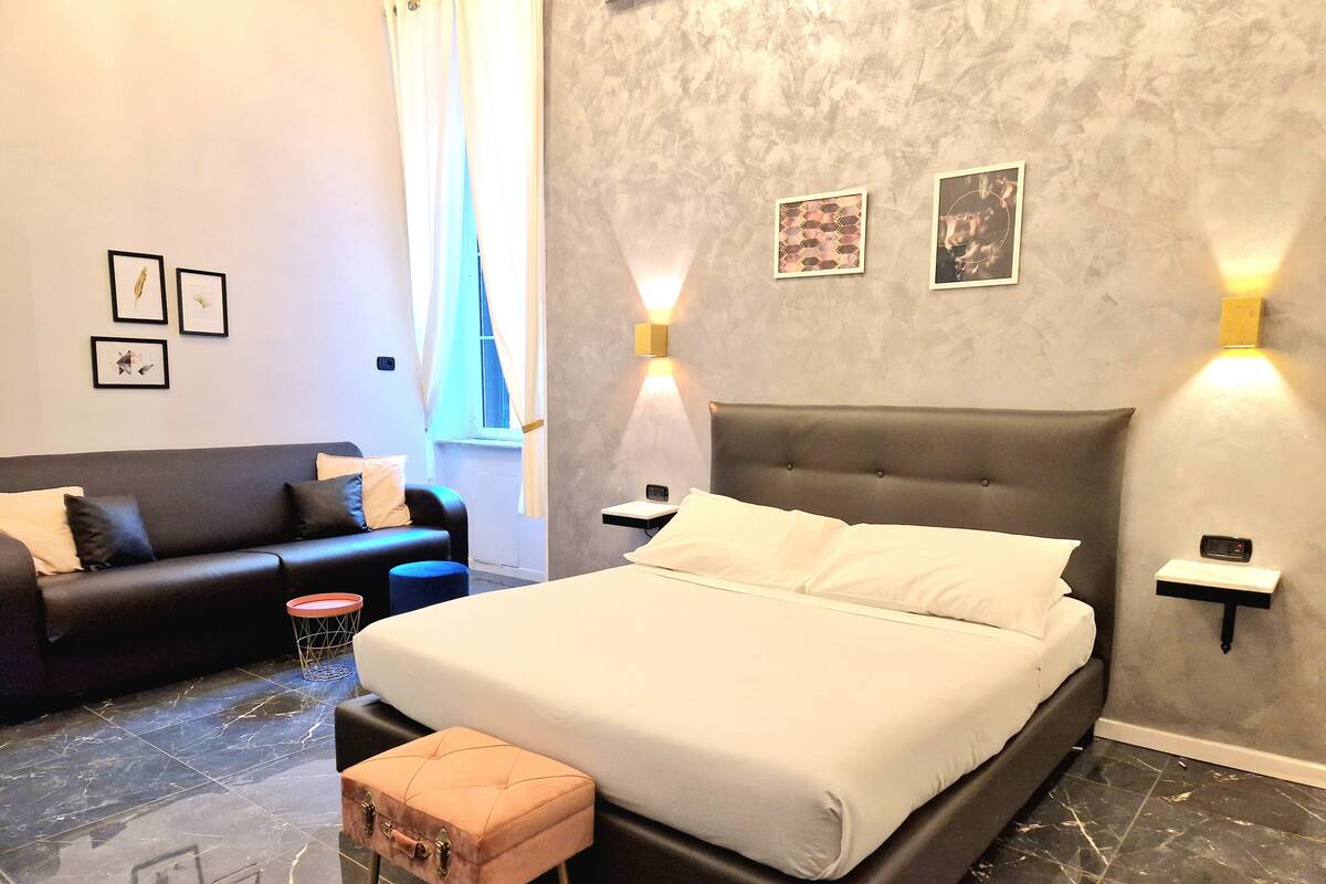 Una habitación de hotel moderna con una cama grande, un sofá y un pequeño taburete. Hay luces fijadas a la pared y cuadros enmarcados.