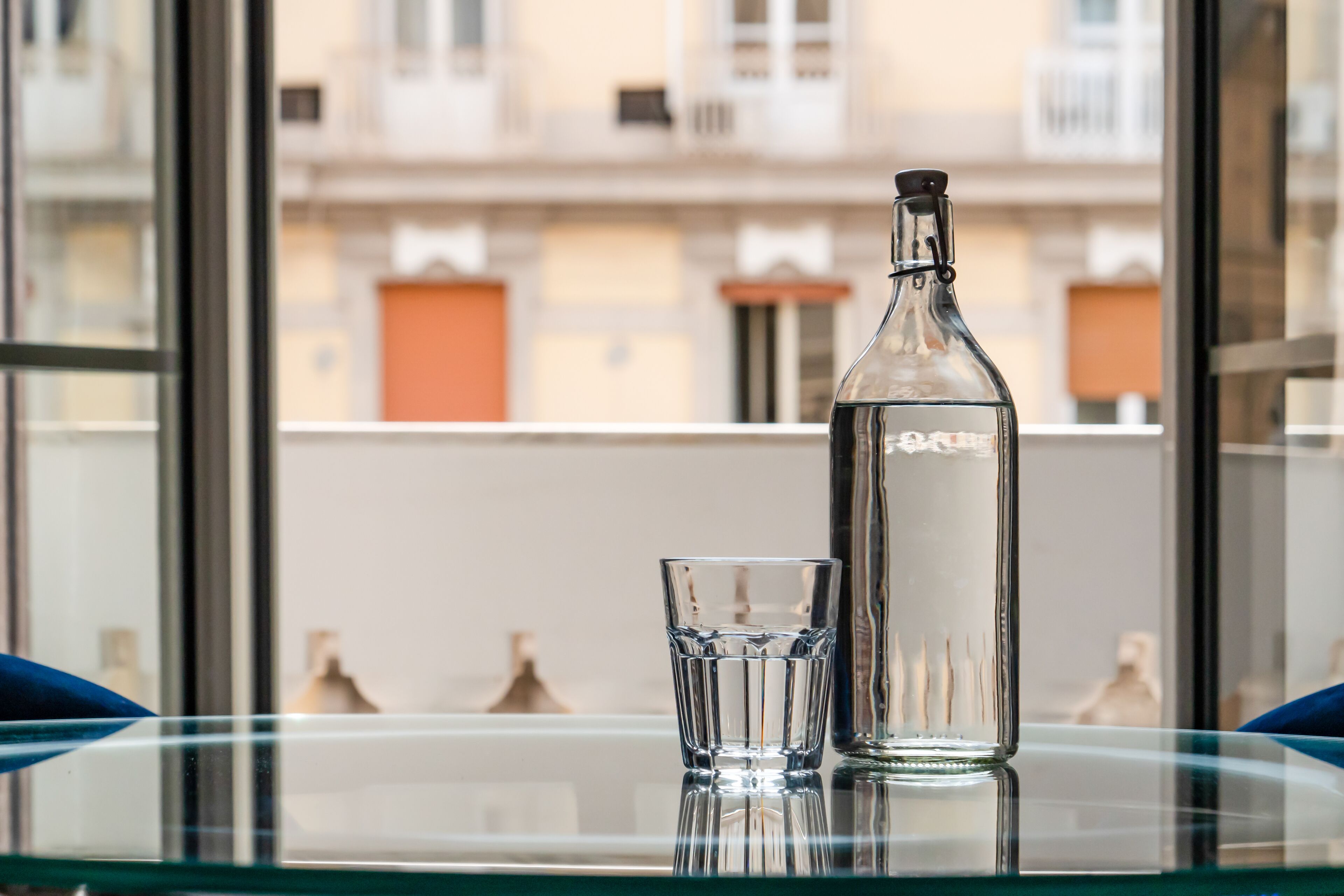 A clear glass bottle and a glass on a reflective surface, with a building in the background.