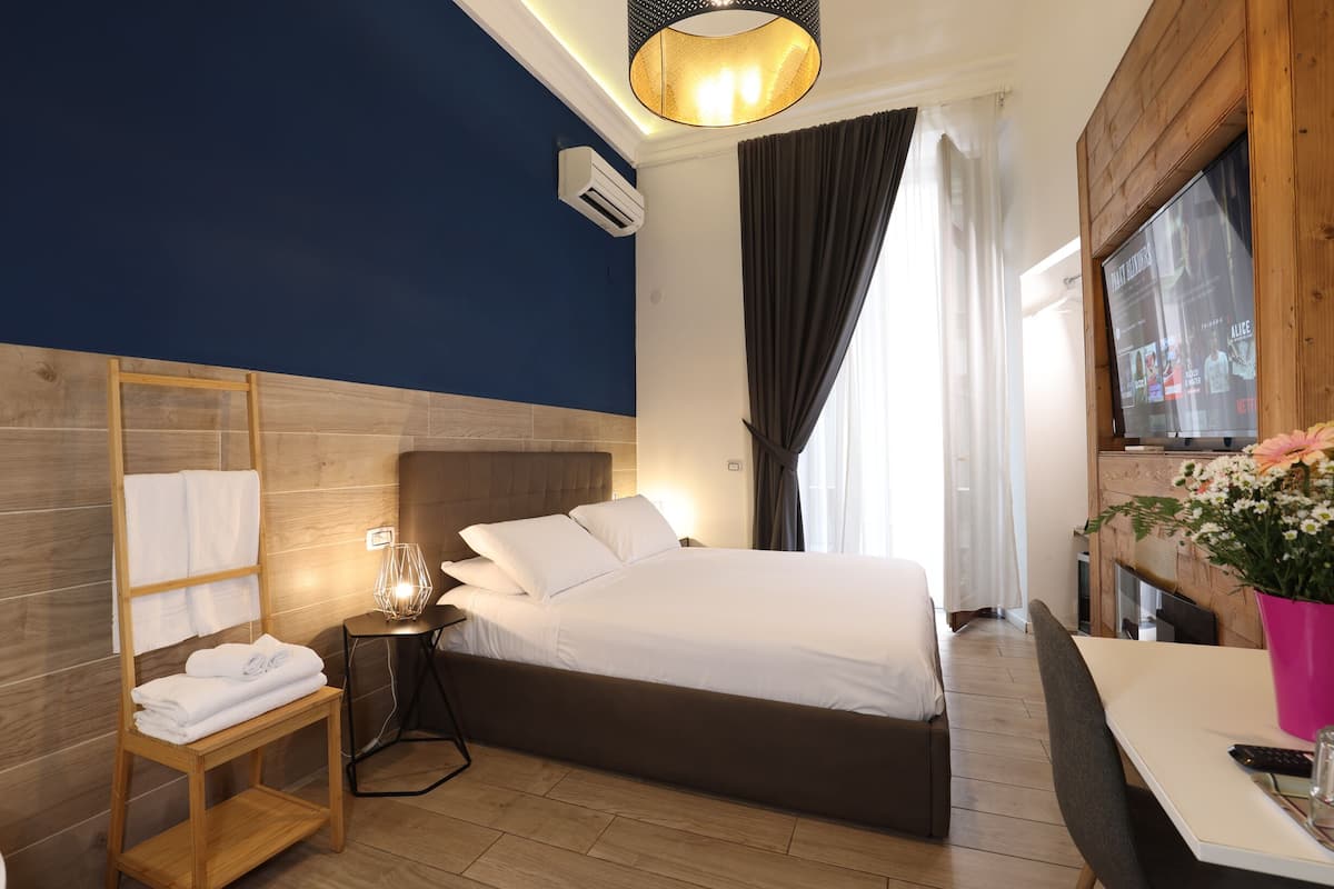 Una habitación de hotel moderna con una cama grande, cabecero de madera, mesita de noche con lámpara, mesita auxiliar de madera con toallas, televisor en una pared de madera y una ventana con cortinas.
