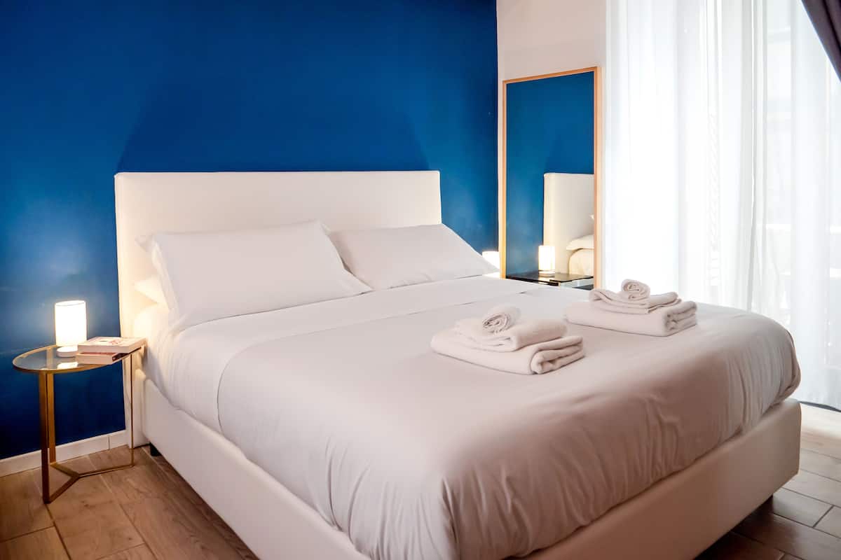 Una cama bien tendida con sábanas y toallas blancas, una mesita de noche con una lámpara y un espejo grande en la pared.