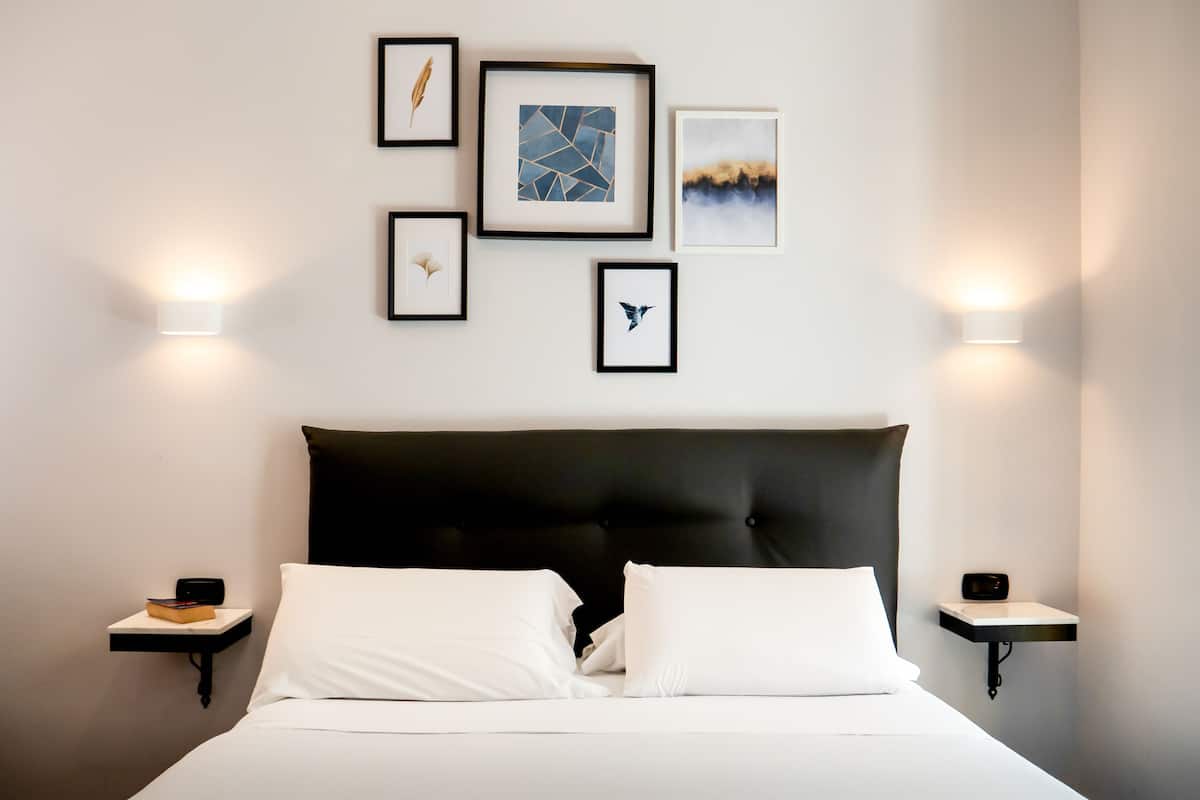 Una cama bien tendida con sábanas blancas y un cabecero negro, flanqueada por dos mesitas de noche con lámparas, y una obra de arte en la pared.