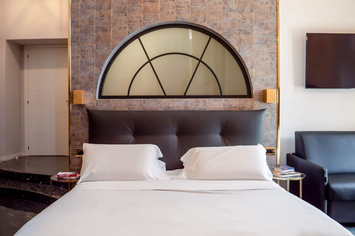 Una habitación de hotel con una cama grande, un ventanal moderno de arco, un televisor de pantalla plana y un sillón de cuero.