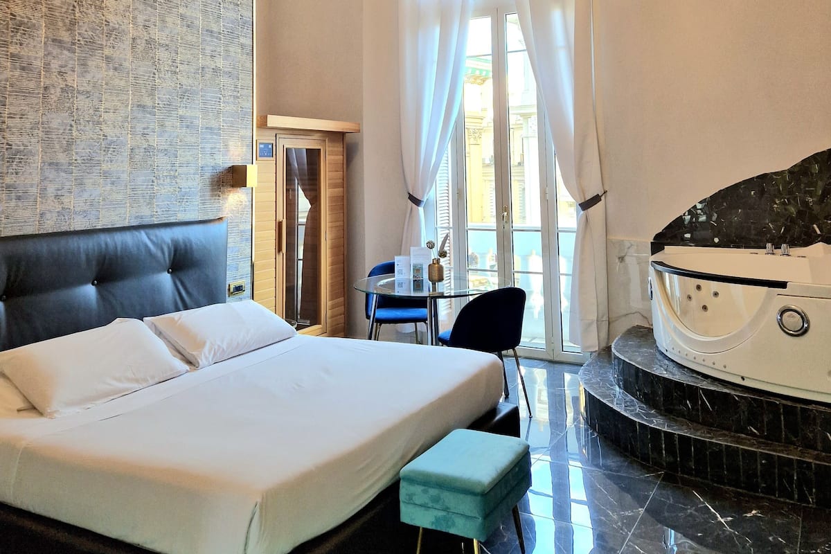 Una habitación de hotel con una cama, una mesita con sillas, una bañera y un ventanal con cortinas.