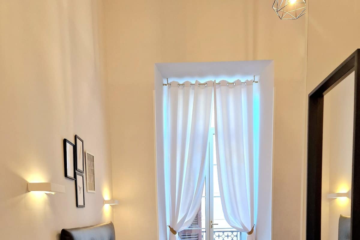 Un dormitorio con cama, mesita de noche, espejo y ventana con cortinas.