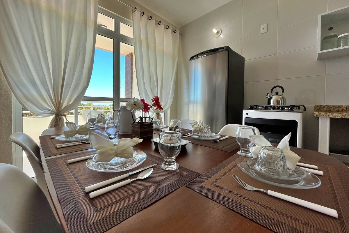 A dining table set with glassware, plates, and cutlery, a kettle, and a view of the outdoors through large windows.