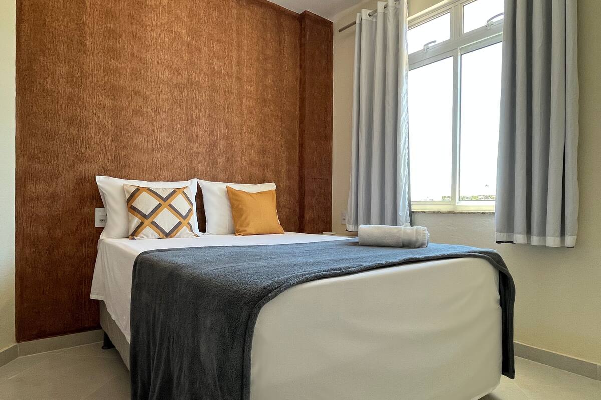 A neatly made bed with a white headboard, a dark blue blanket, and two pillows, one with a geometric pattern and the other solid orange.