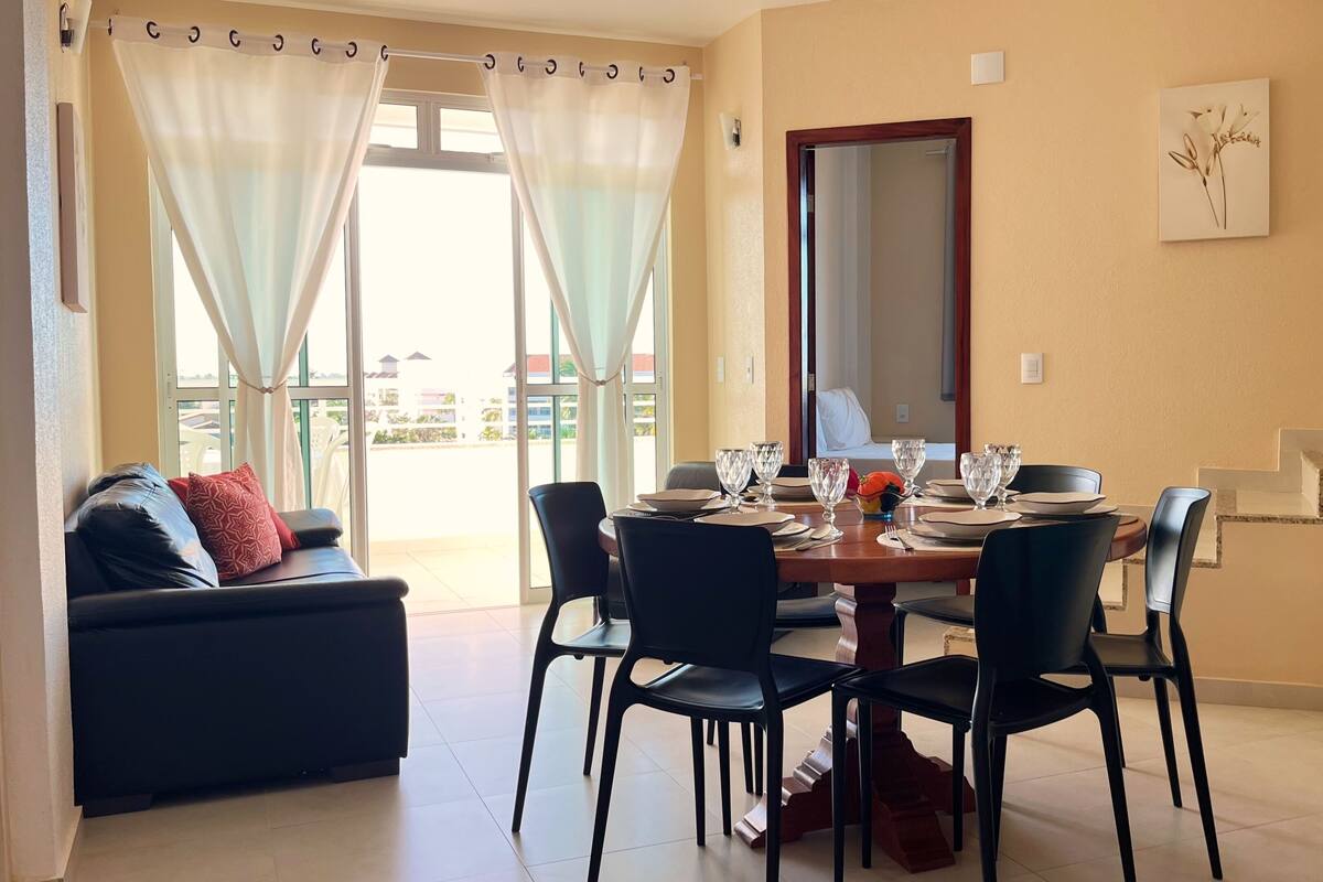 A dining area with a table set for four, a sofa with cushions, and a view of the outside through large windows.