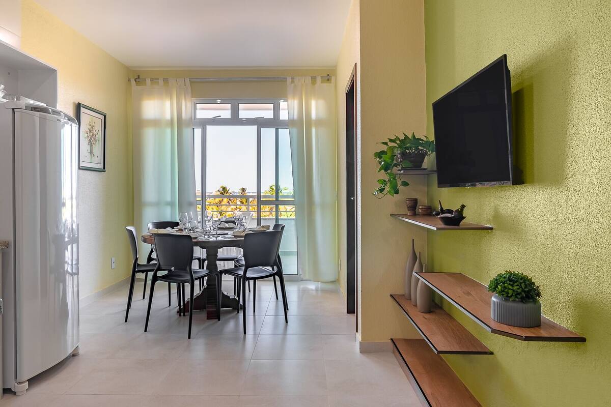 A dining area with a table set for four, a television mounted on the wall, and a balcony with a view.