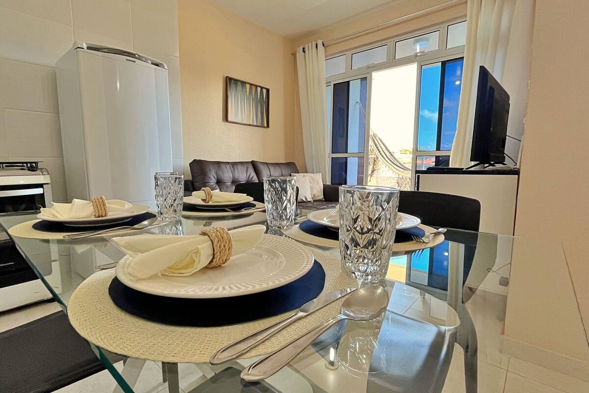 A dining area with a glass table set for four, a sofa in the background, and a view of the outside through a window.