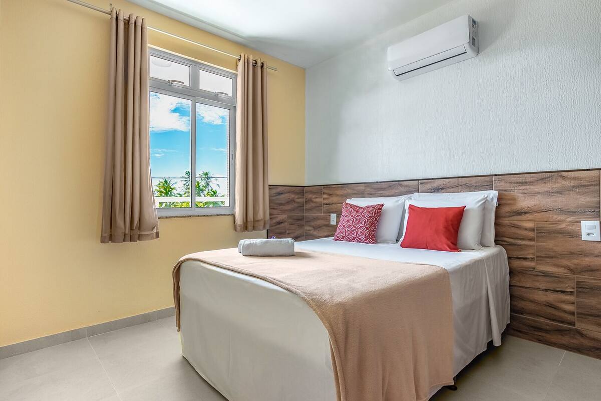 A hotel room with a bed, curtains, an air conditioning unit, and a view of greenery outside.