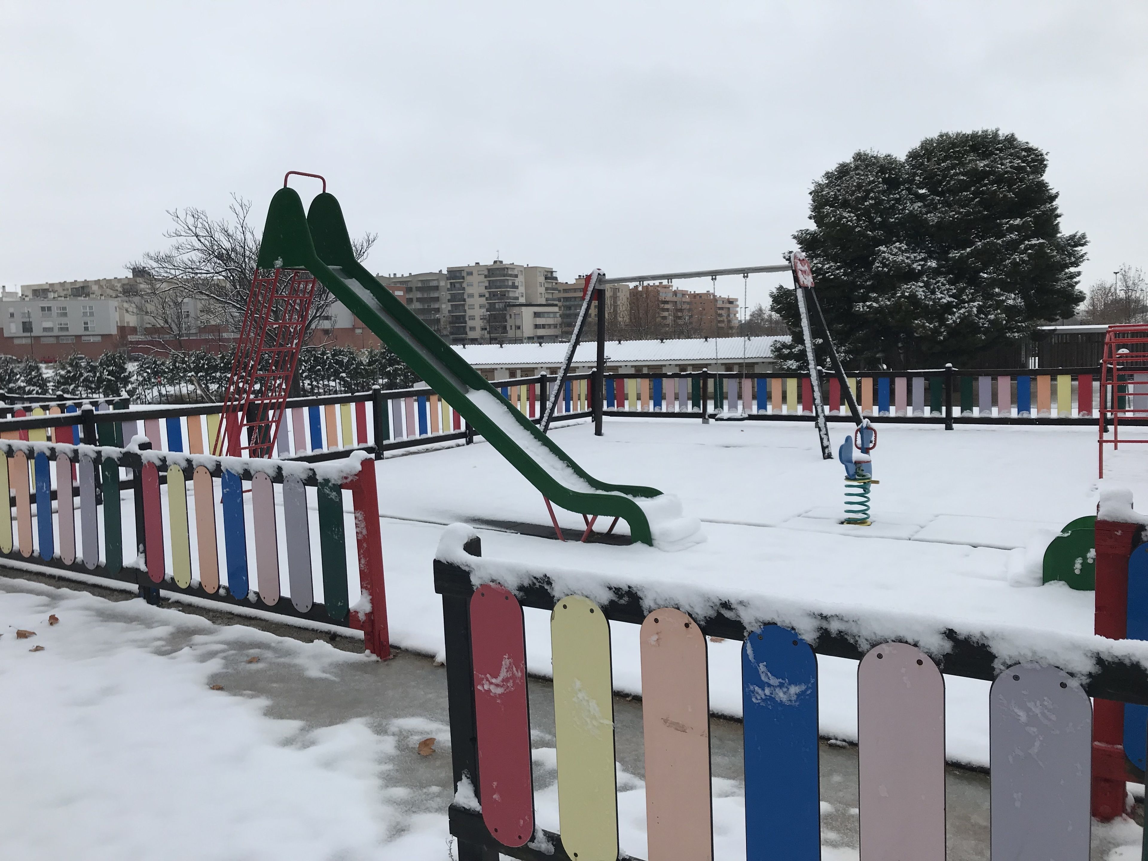 A playground with a green slide and a red climbing structure, surrounded by a fence with colorful panels.