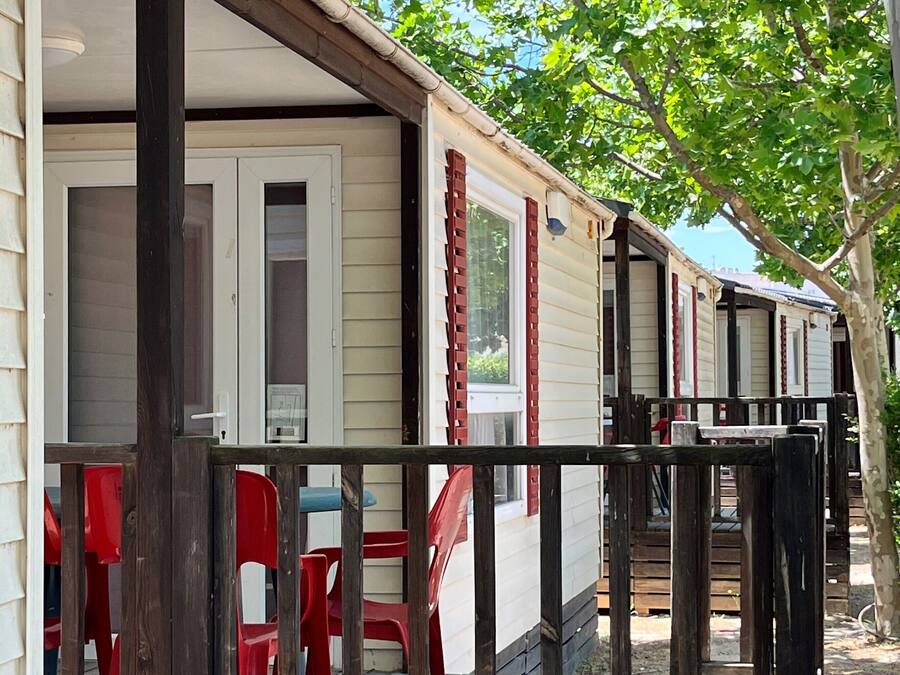 Une rangée de maisons mobiles dotées d’une terrasse en bois et de chaises rouges, entourées d’arbres.