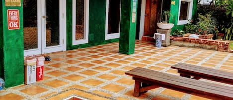 A green building with a tiled patio, a wooden bench, and a trash bin.
