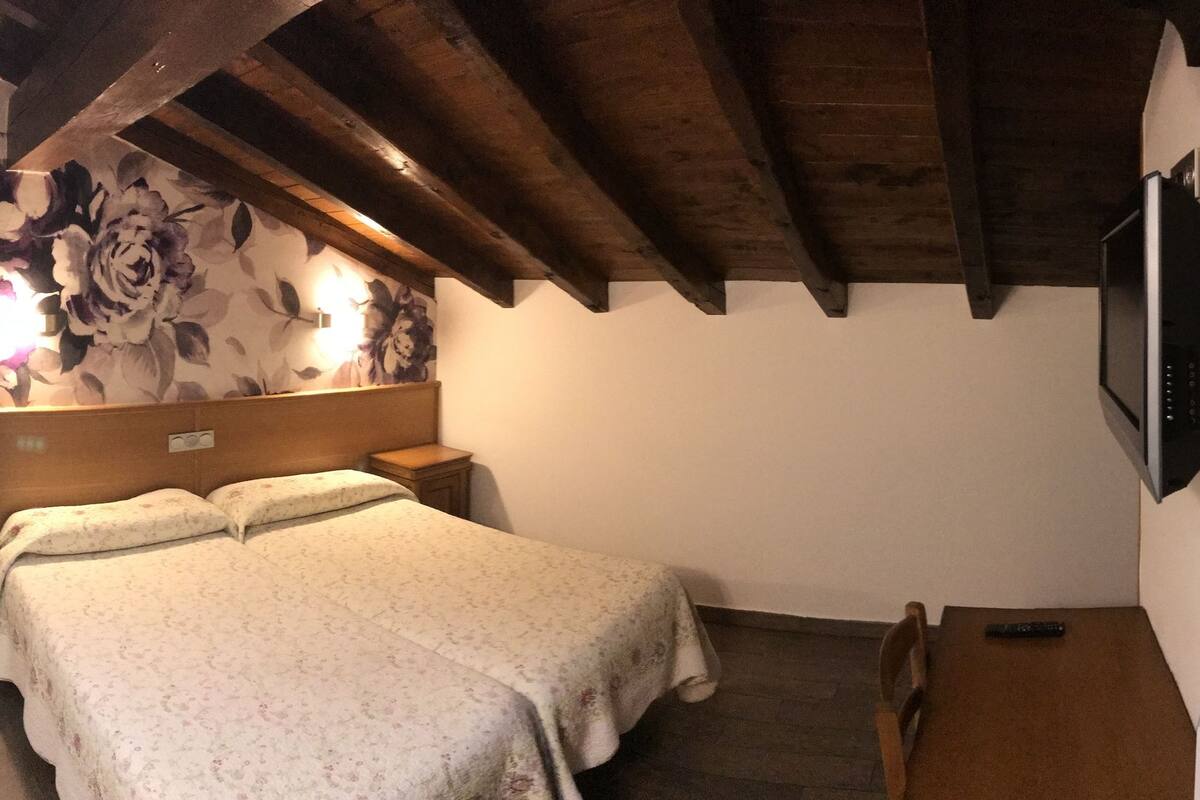 A bedroom with a bed, a wooden ceiling, a wall-mounted TV, and a bathroom with a sink and mirror.
