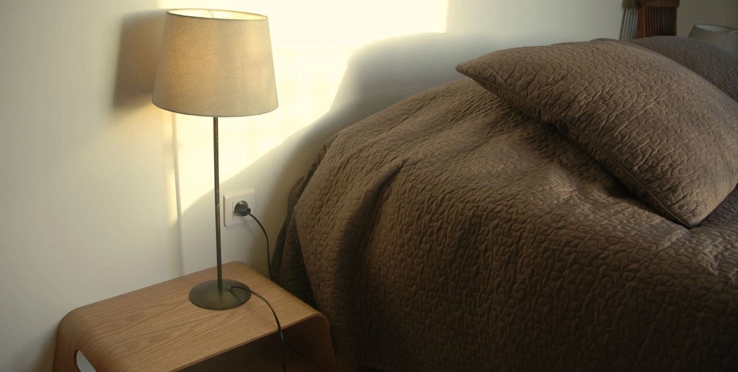 Un lit recouvert d’une couverture à motifs, une lampe de chevet posée sur une table de nuit en bois et une prise électrique au mur.