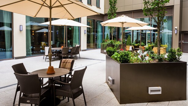 An outdoor patio with round tables and chairs, large umbrellas, and a planter box with greenery. - Maldron Hotel Newcastle (Newcastle-upon-Tyne)