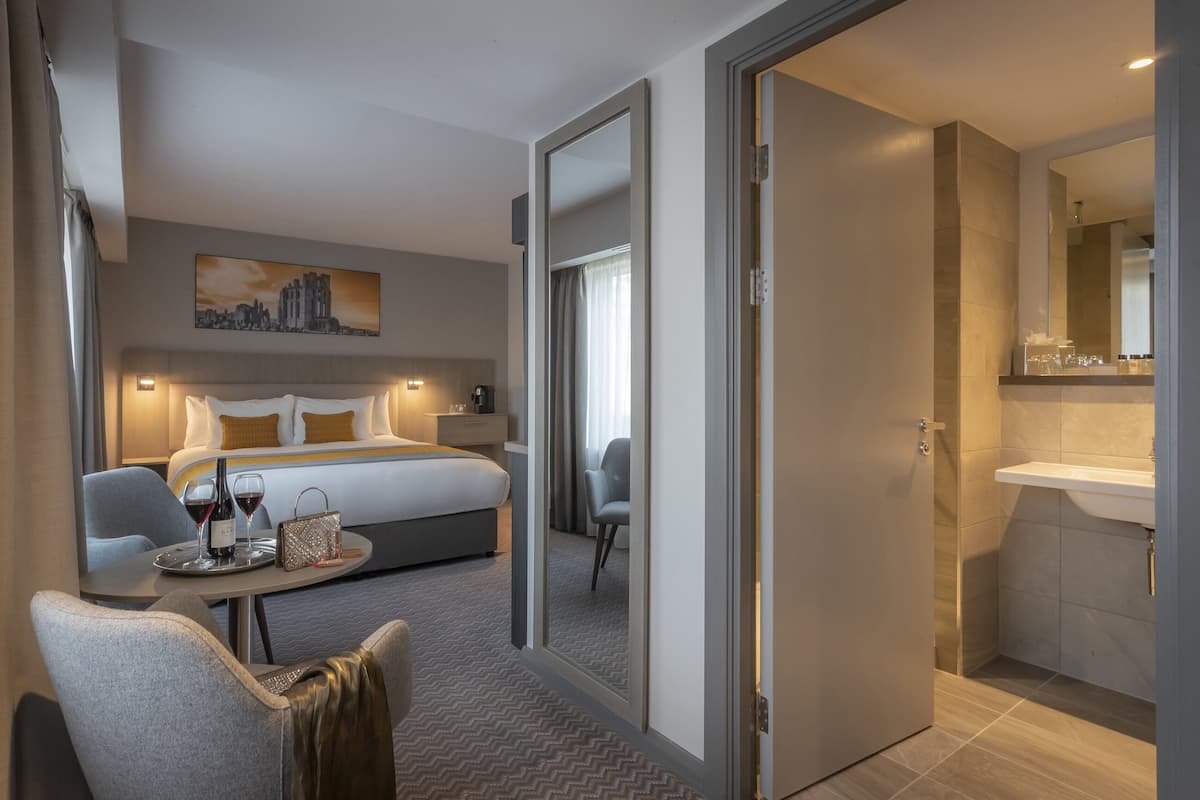 A hotel room with a bed, a desk with a chair, a small table with wine glasses and a bottle, and a bathroom with a sink and mirror.