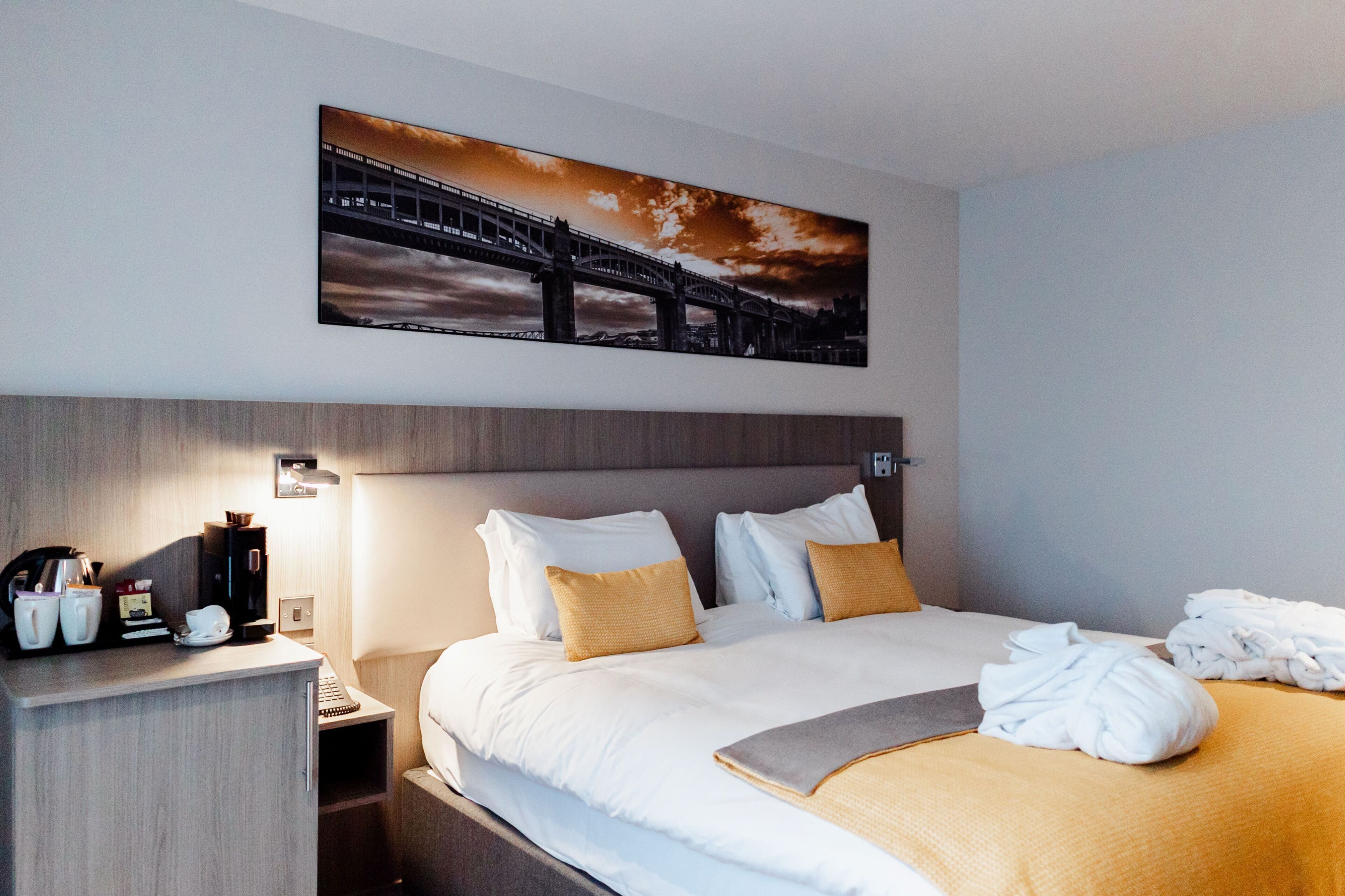 A hotel room with a bed, pillows, a nightstand with a coffee maker, and a painting of a bridge.