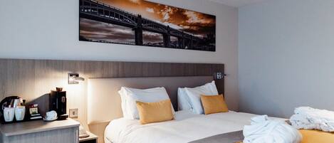 A hotel room with a bed, pillows, a nightstand with a coffee maker, and a painting of a bridge.