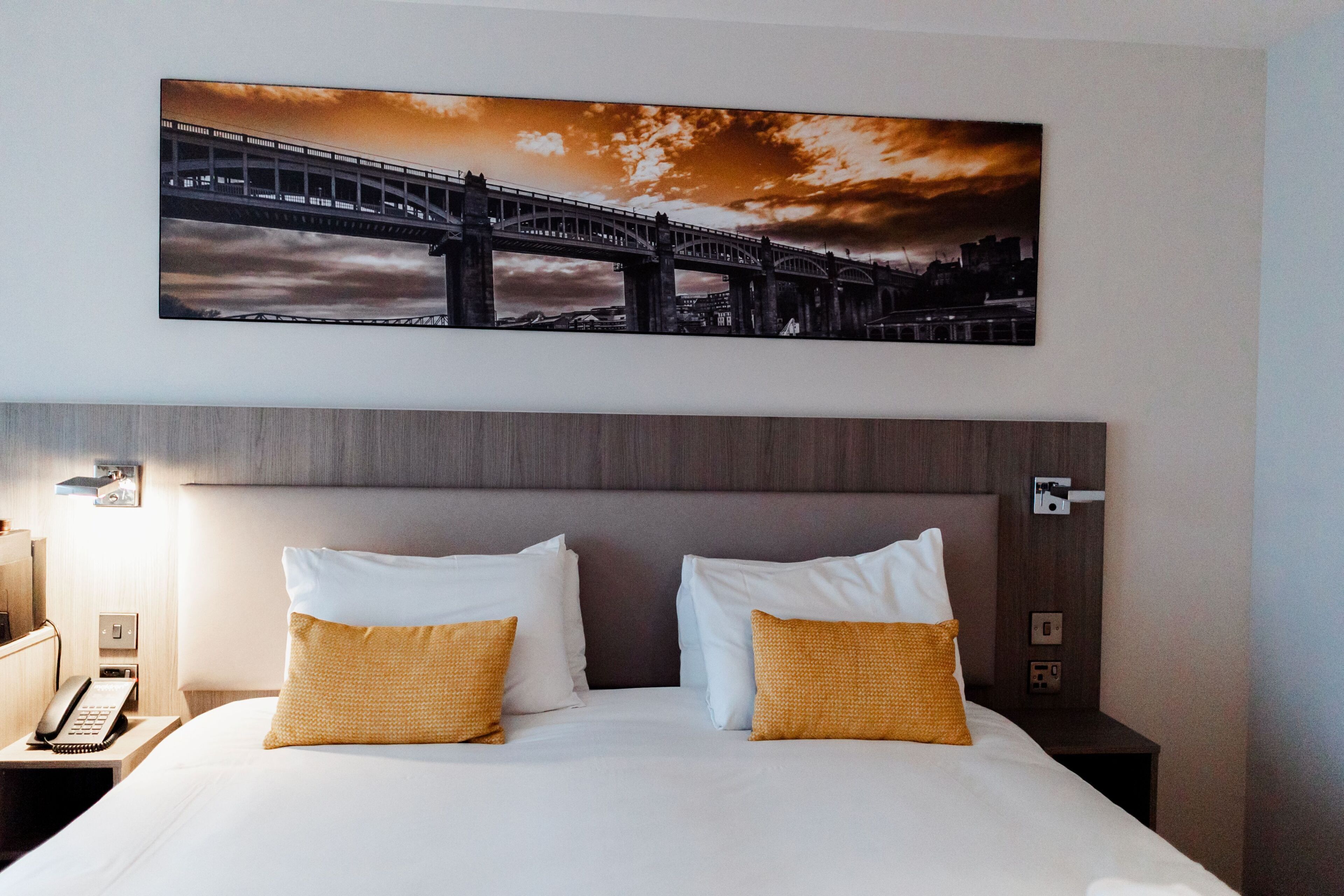 A hotel room with a bed, two pillows, a nightstand with a phone, and a wall-mounted picture of a bridge.