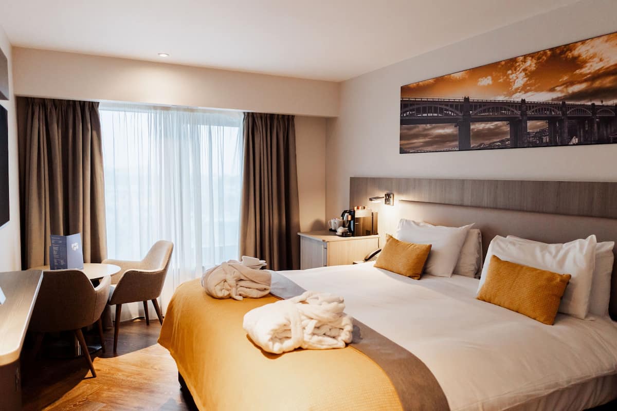 A hotel room with a bed, a desk, chairs, a nightstand, and a painting on the wall.