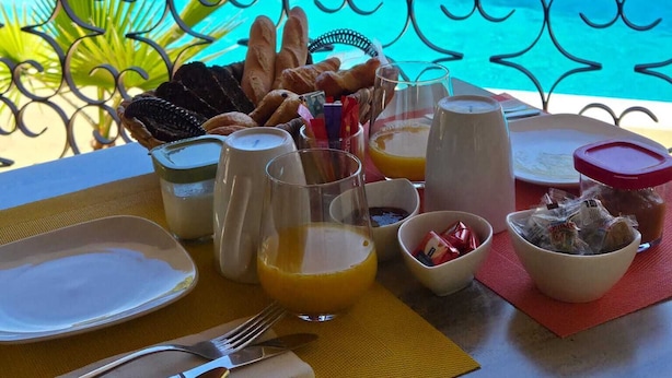 A table set with breakfast items, a glass of orange juice, and a view of a pool and greenery. - Villa Manoé (Draguignan)