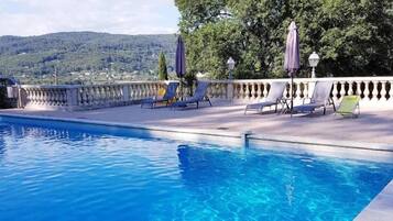 A swimming pool with clear blue water, surrounded by lounge chairs and umbrellas, with a view of trees and hills in the background.