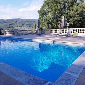 A swimming pool with clear blue water, surrounded by lounge chairs and umbrellas, with a view of trees and hills in the background. - Villa Manoé (Draguignan)