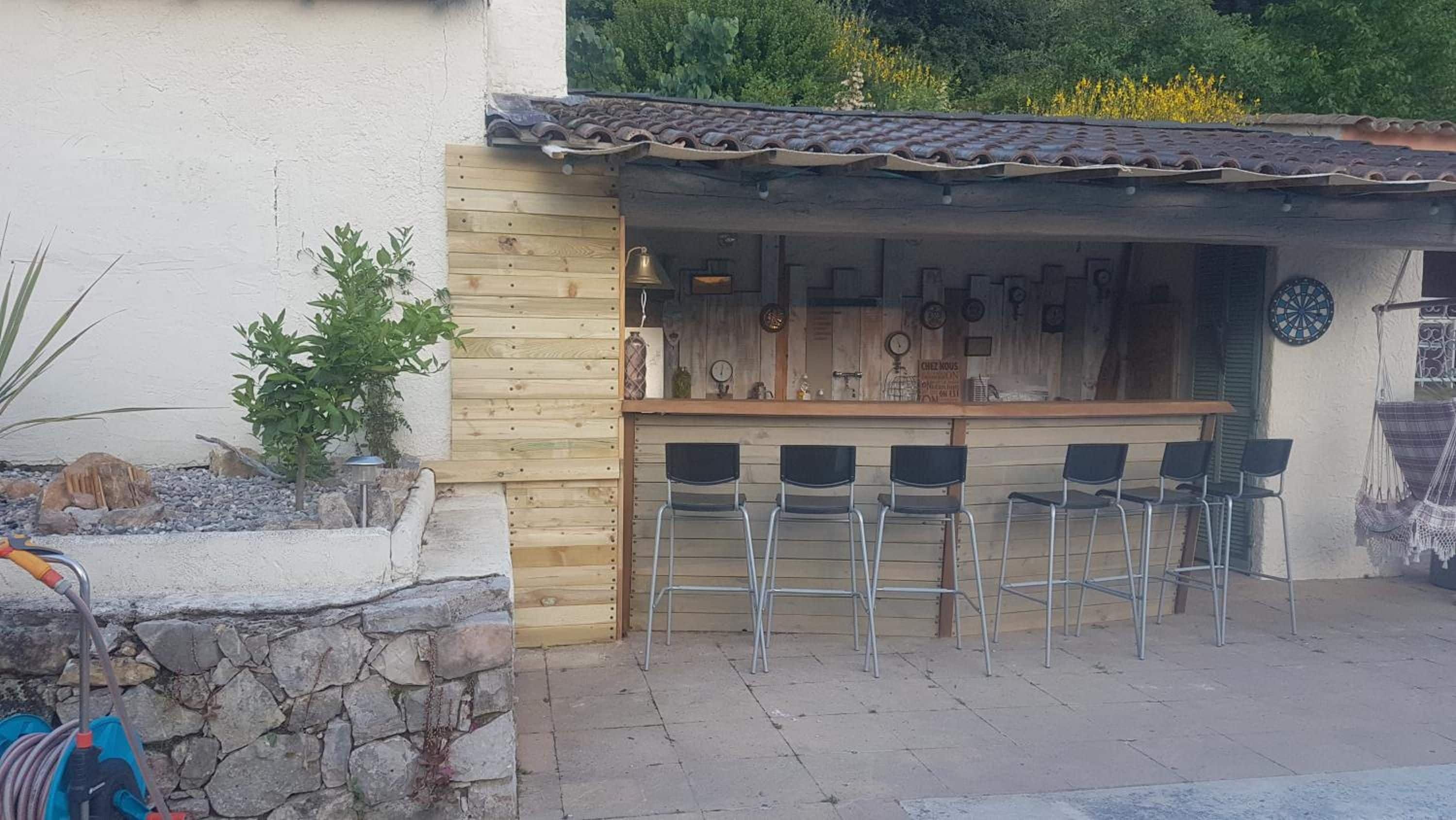 A rustic outdoor bar area with wooden walls, metal bar stools, and a tiled roof.