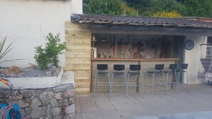 A rustic outdoor bar area with wooden walls, metal bar stools, and a tiled roof. - Villa Manoé (Draguignan)