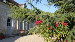 Un edificio de piedra con camino de grava y jardín con arbustos verdes y flores rosadas.