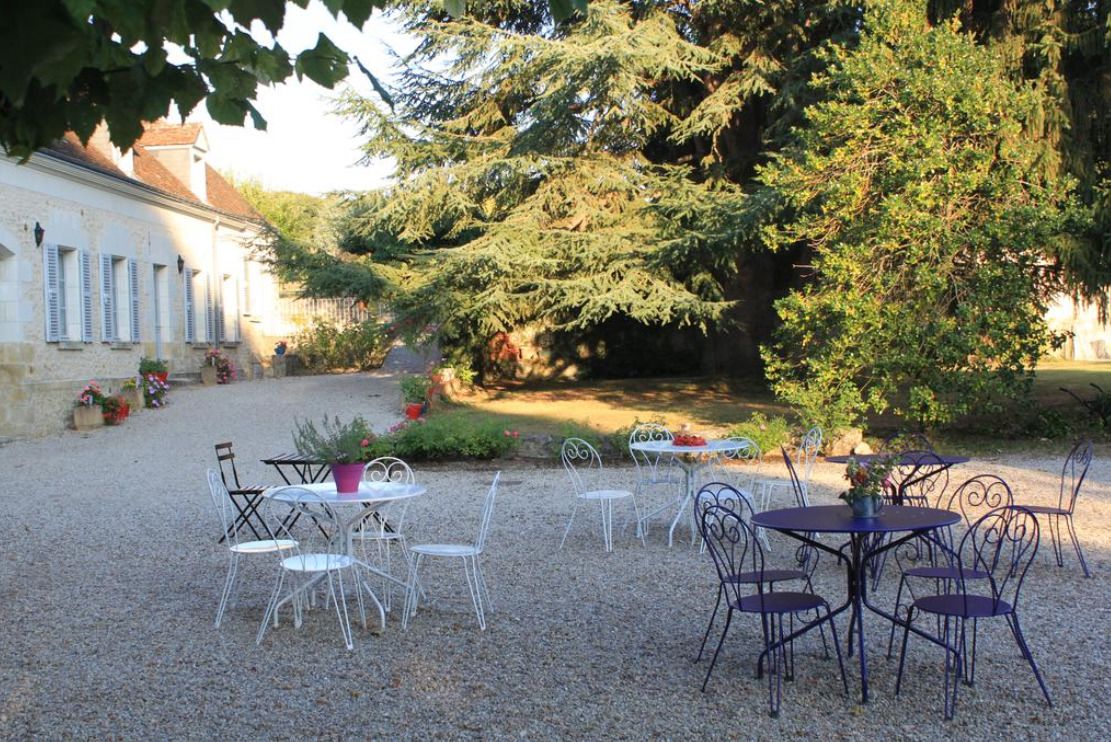 An outdoor seating area with tables and chairs, surrounded by gravel and plants, with a building in the background.