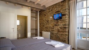 A bedroom with a stone wall, a television mounted on the wall, a bed with a grey bedspread, a white bathroom with a towel on the bed, and a window with curtains. - Pensión Domus Gallery (Arzua)