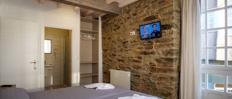 A bedroom with a stone wall, a television mounted on the wall, a bed with a grey bedspread, a white bathroom with a towel on the bed, and a window with curtains.