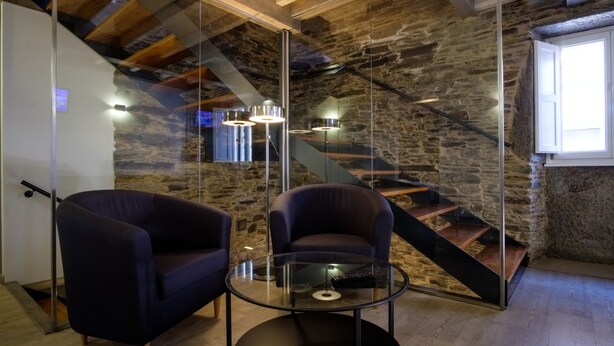 A modern interior with a stone wall, a staircase, and two dark armchairs. - Pensión Domus Gallery (Arzua)