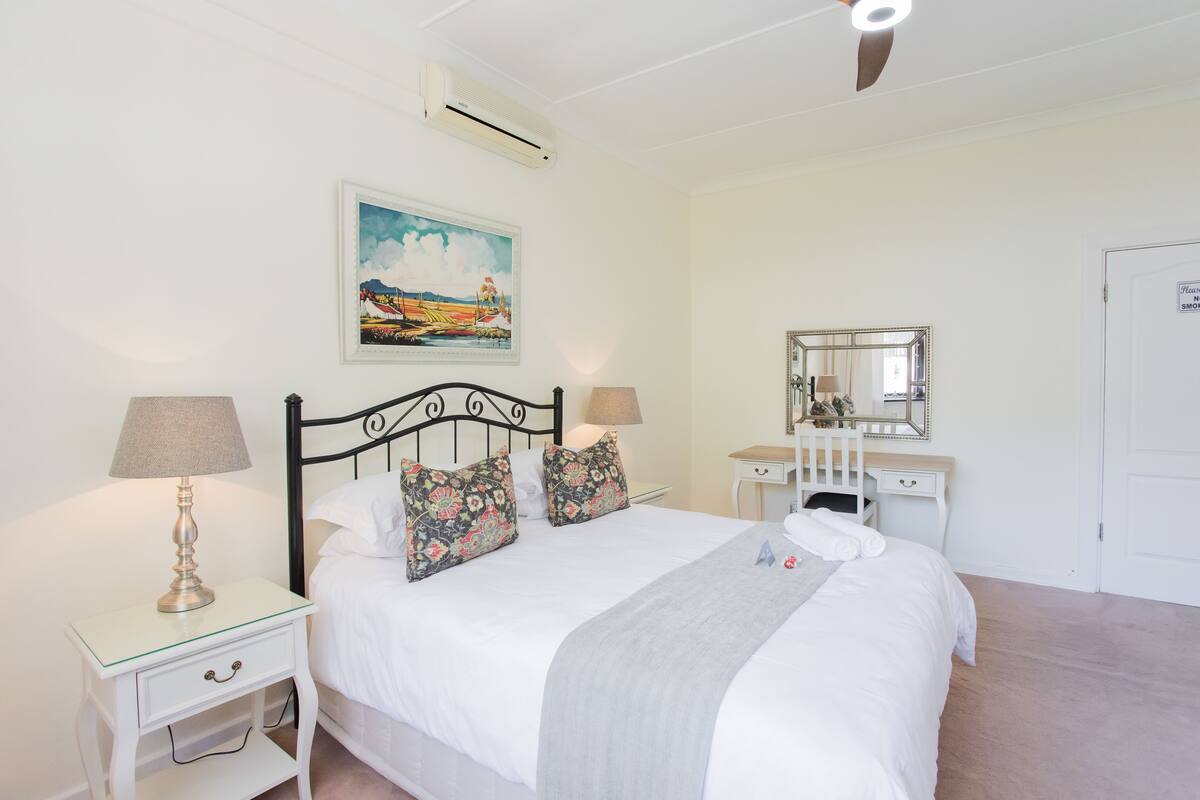 A neatly arranged bedroom with a bed, bedside tables, a dresser, a mirror, and a painting on the wall.