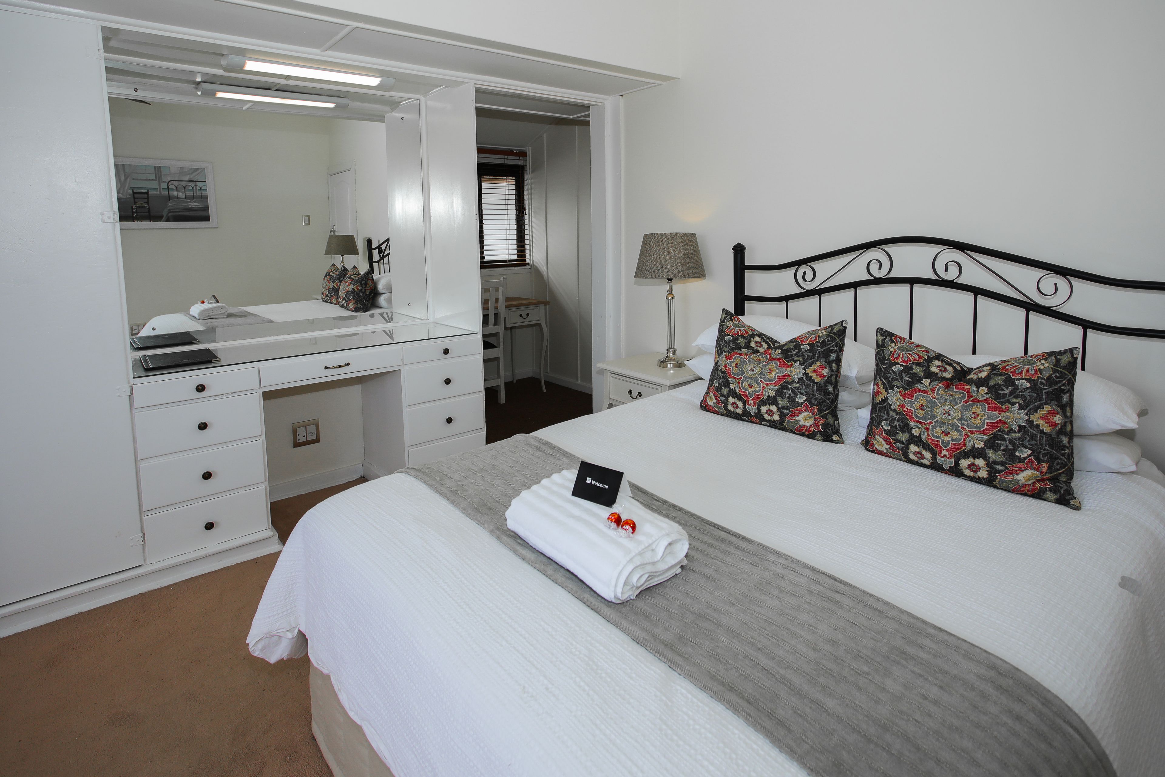 A neatly arranged bedroom with a bed, a bedside table with a lamp, a dresser, and a mirror.