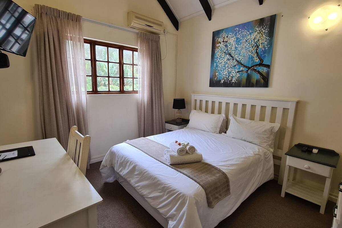 A hotel room with a bed, a desk, a chair, a TV, a painting, and a window with curtains.