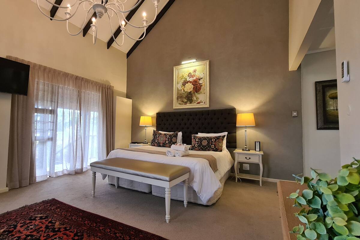 A bedroom with a large bed, two bedside tables, a chandelier, a painting, and a potted plant.