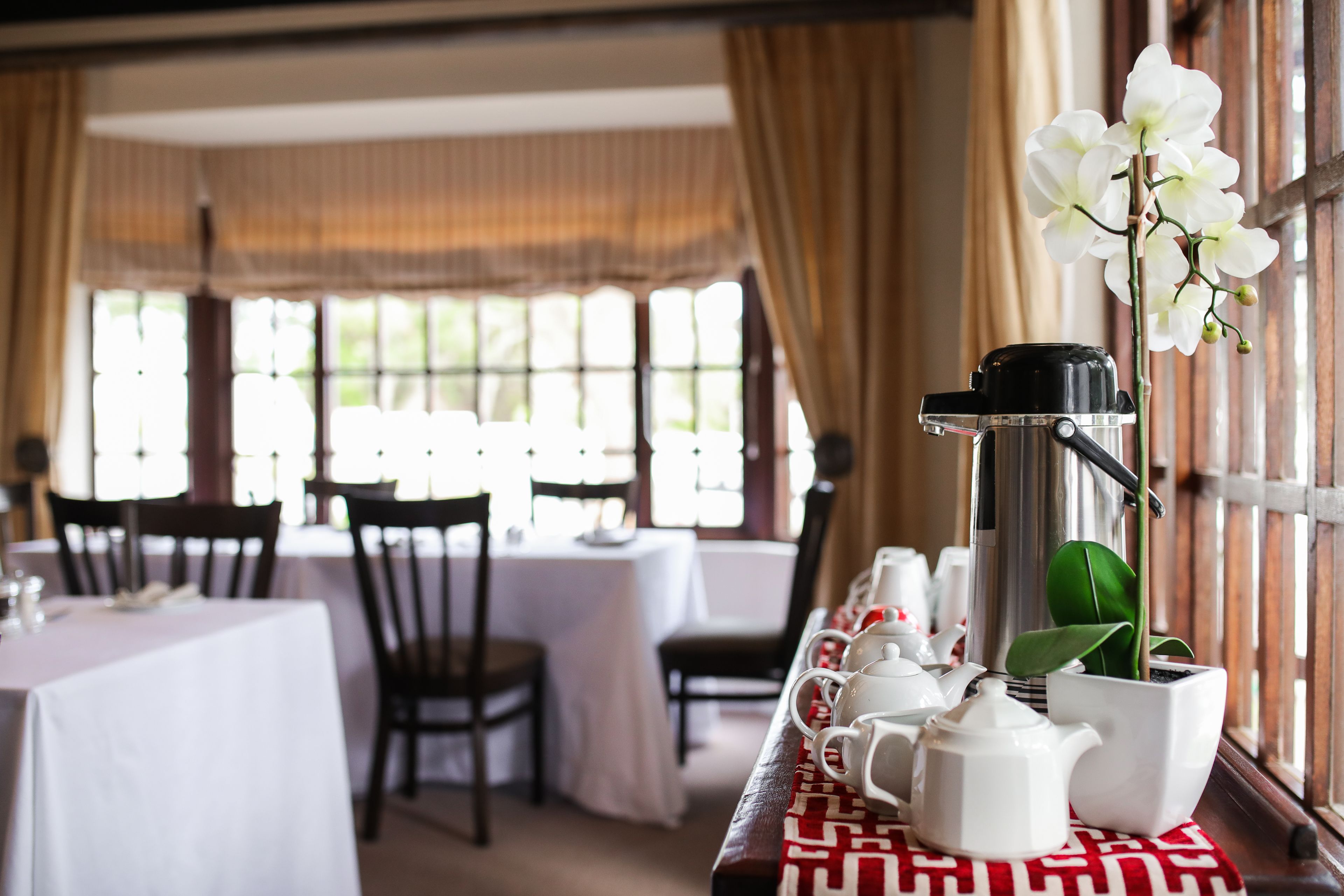 Un comedor con mesas dispuestas para una comida, una cafetera y una orquídea en maceta.