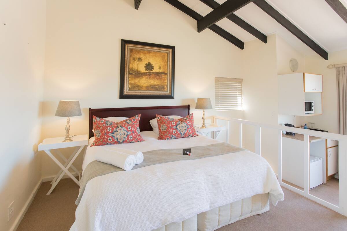 A bedroom with a bed, two bedside tables, a desk, a chair, a lamp, a painting, and a window with blinds.