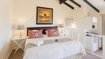 A bedroom with a bed, two bedside tables, a desk, a chair, a lamp, a painting, and a window with blinds.