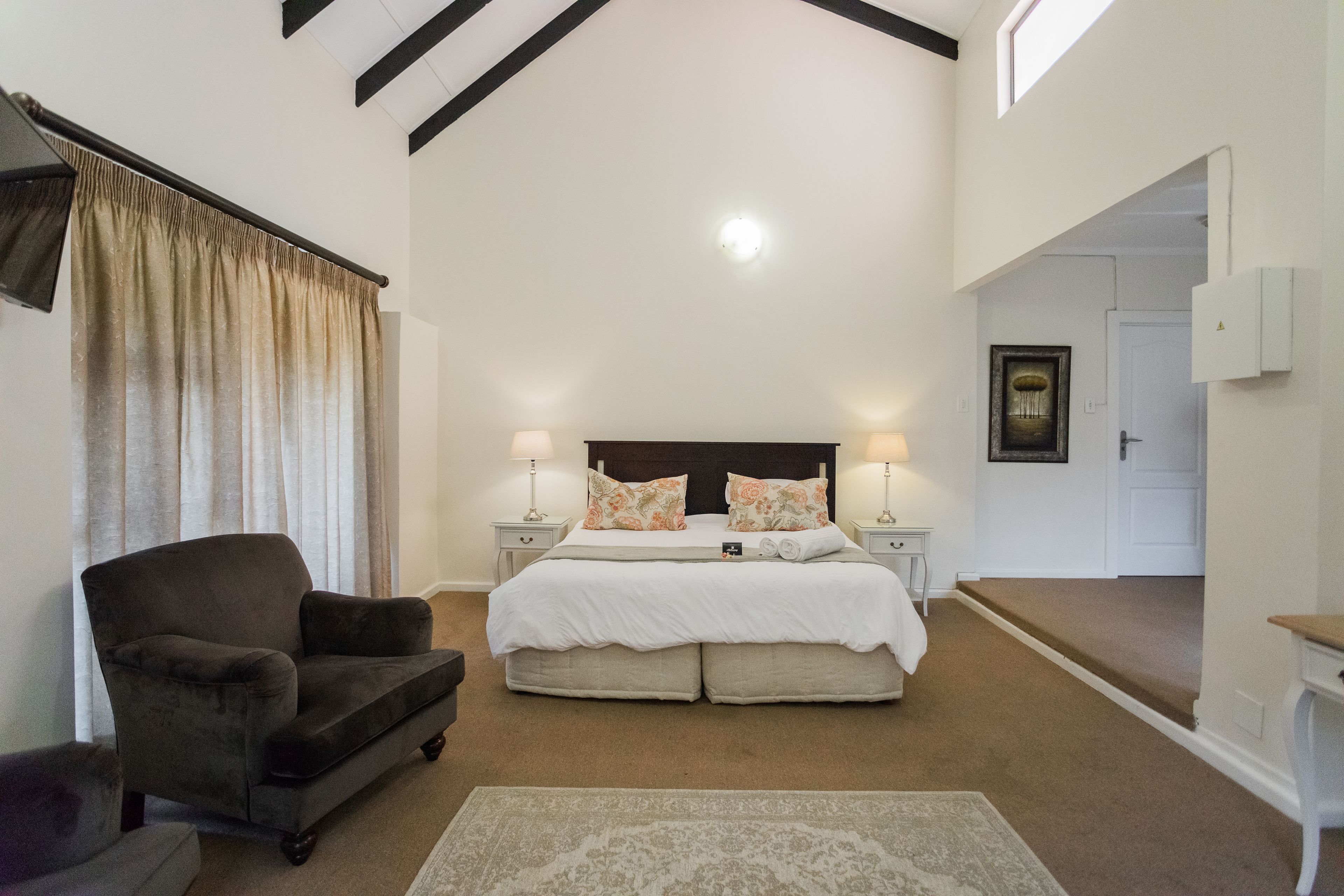 A bedroom with a large bed, two bedside tables with lamps, a dark armchair, and a large window with curtains.