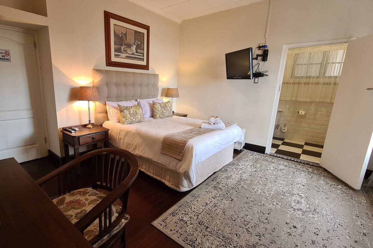 A hotel room with a bed, two bedside lamps, a television mounted on the wall, a door with a 'No Smoking' sign, a chair, and a small table with a plant.