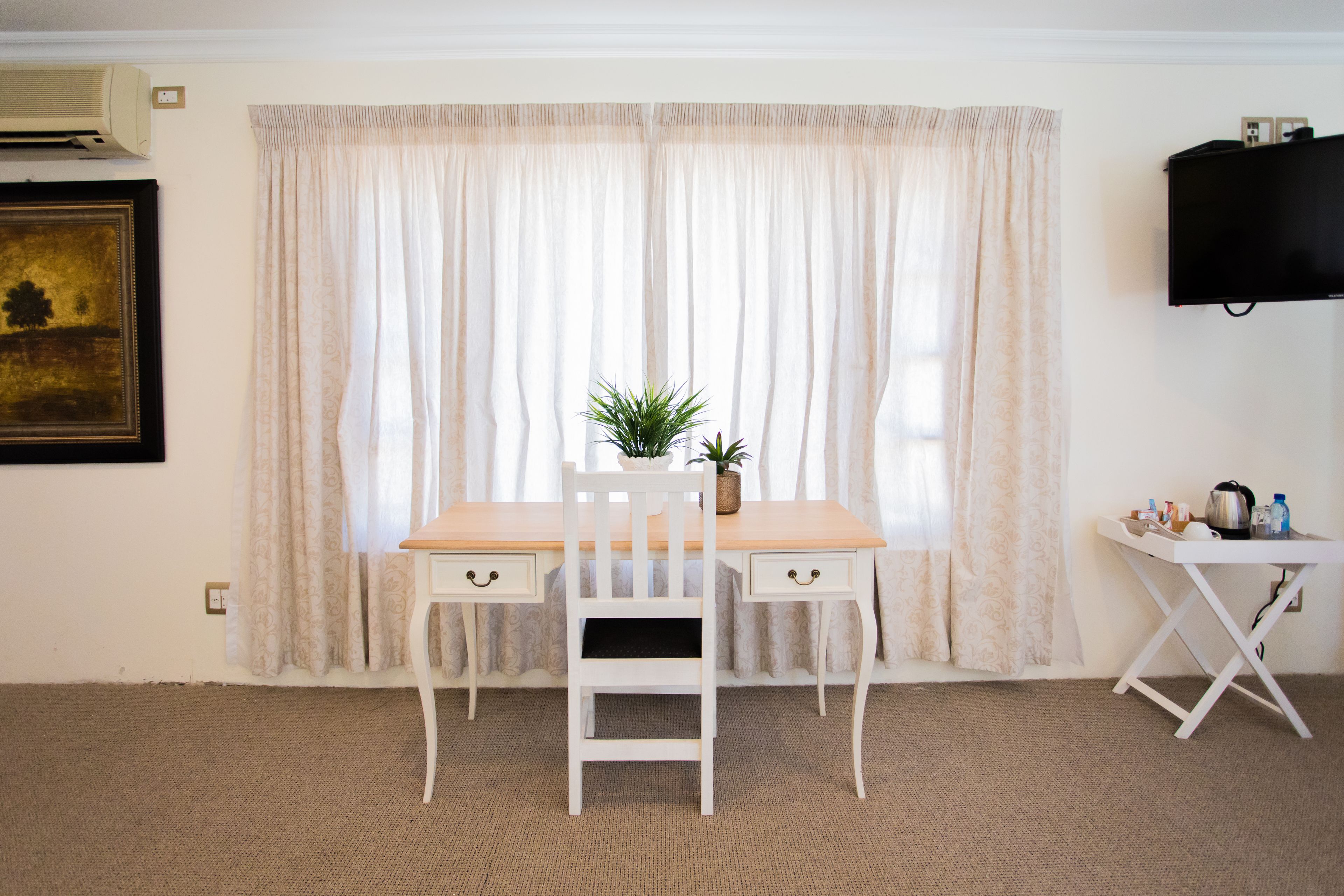 A room with a wooden desk, two chairs, a small table with a plant, and a television on the wall.