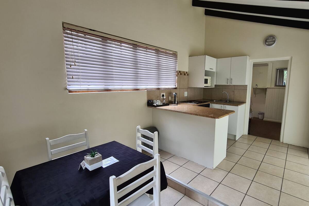 A compact kitchen with a dining area, featuring a small table, chairs, a window with blinds, and a view into the adjacent room.