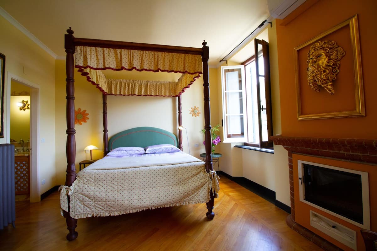Uma cama com dossel com colcha florida, mesa de cabeceira com abajur e lareira com moldura decorativa.