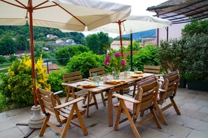 A wooden table set outdoors with chairs, a white umbrella, and a view of a hillside with buildings.