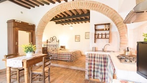 Une pièce rustique avec une arche en pierre, une table en bois entourée de chaises, un lit avec une literie à carreaux, un coin cuisine avec une cuisinière à gaz et un luminaire suspendu.