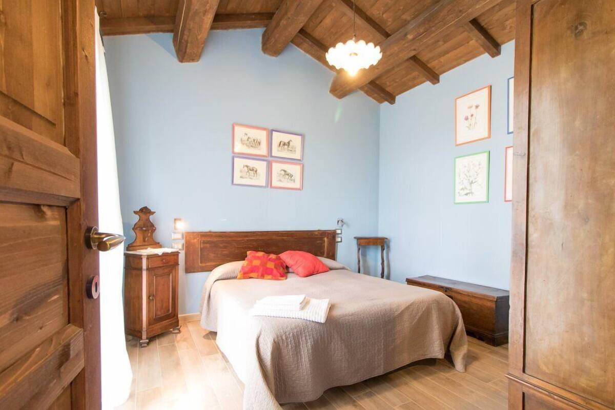 Een slaapkamer met een houten plafond, een bed met een beige dekbedovertrek, een nachtkastje met een lamp en ingelijste foto’s aan de muur.